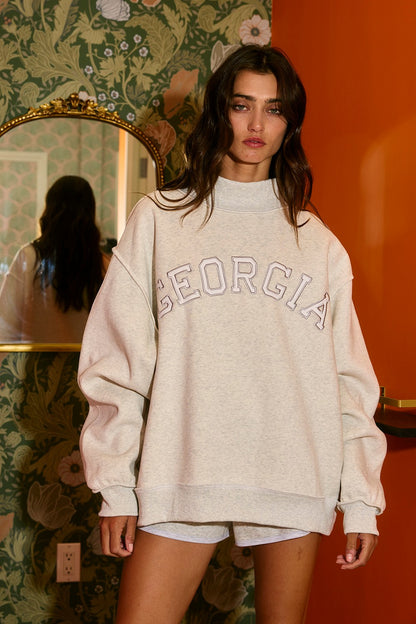 Woman wearing a beige 'Georgia' sweatshirt in a room with floral wallpaper and an orange wall.