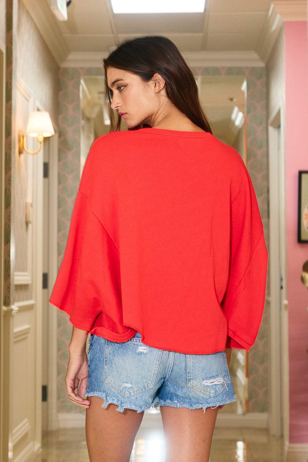 Woman wearing a red top and denim shorts in a hallway.