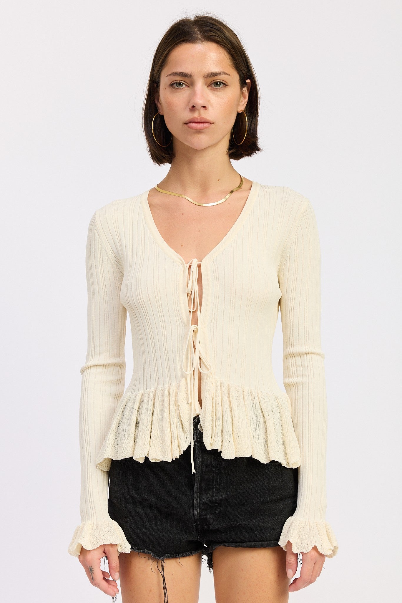 Model wearing a cream ribbed long sleeve top with front tie detail and ruffle peplum hem