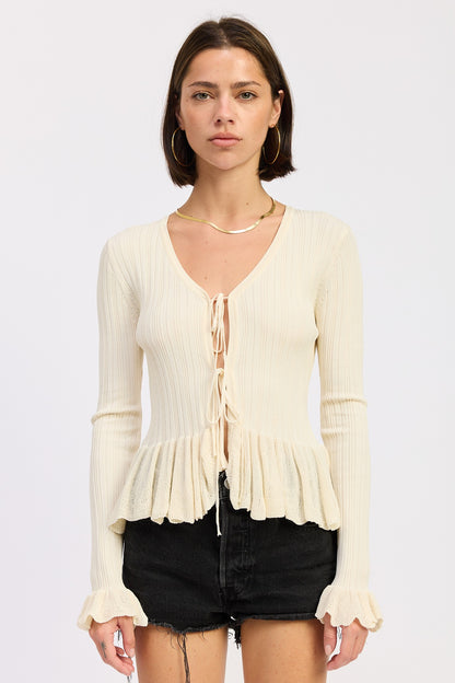 Model wearing a cream ribbed long sleeve top with front tie detail and ruffle peplum hem