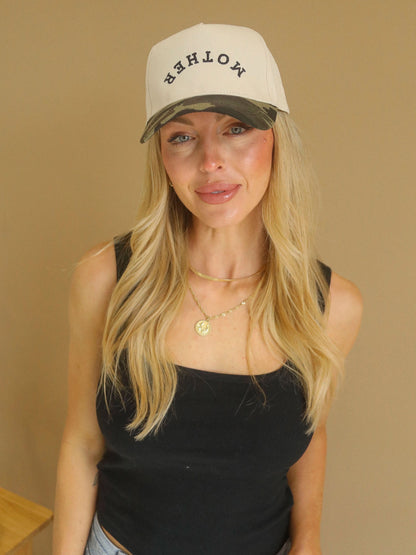Woman wearing a beige and camo two-tone trucker hat with upside-down embroidered “mother” text styled with a black tank and gold jewelry.