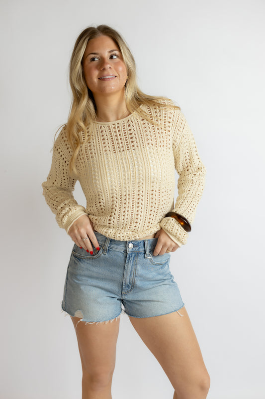 Woman wearing a beige crochet sweater and denim shorts on a plain background