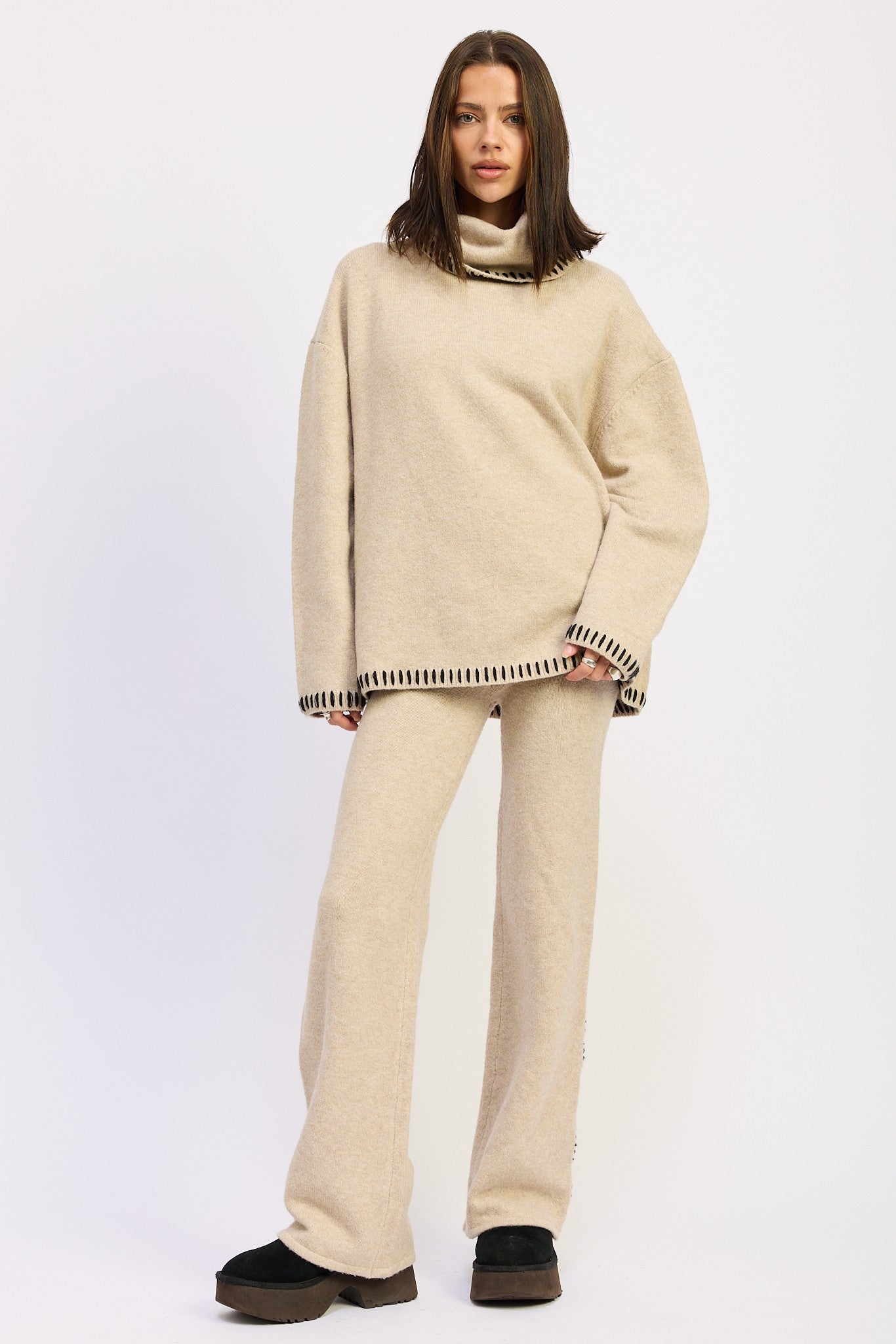 Woman wearing beige wide-leg knit pants with blanket stitching along the sides paired with a matching oversized turtleneck sweater.