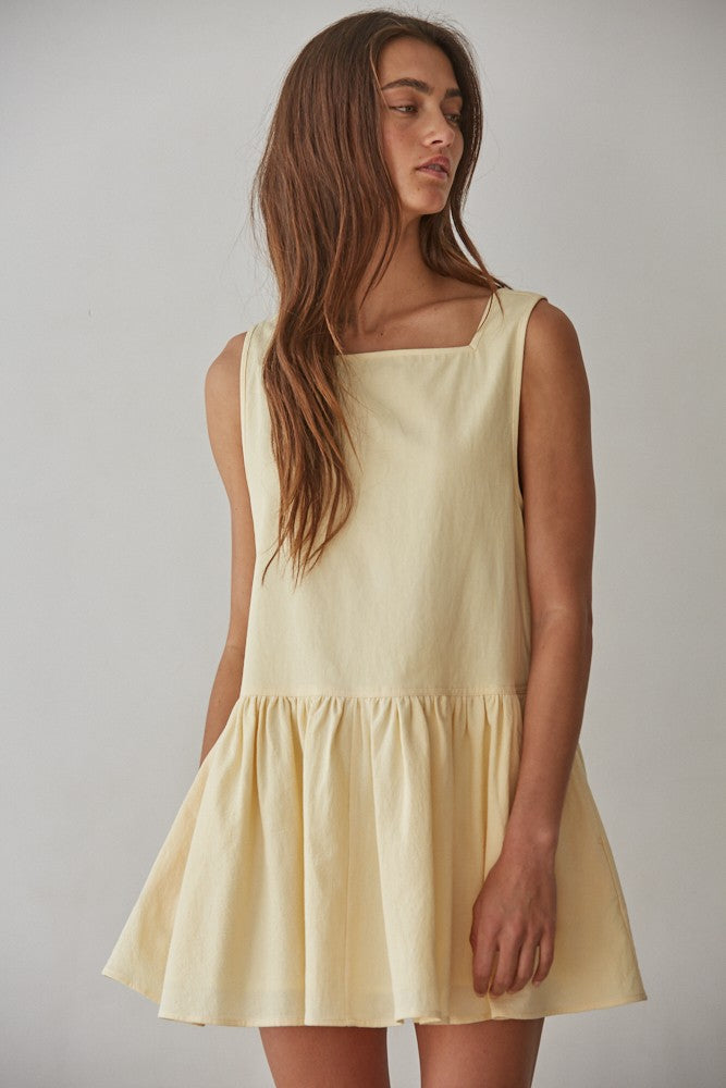 Woman wearing buttercream yellow sleeveless cotton mini dress with square neckline, drop waist flare skirt, and side pockets