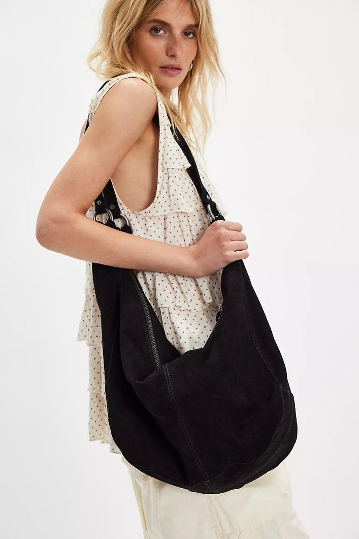 Model carrying black suede hobo tote bag with elongated straps and zipper closure.