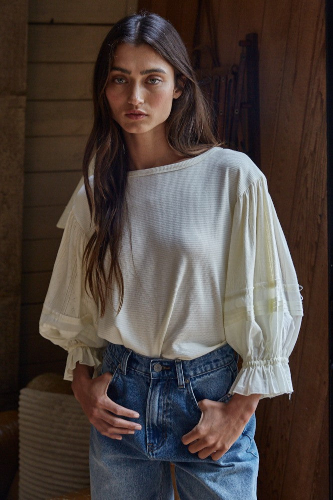 Woman wearing ivory knit puff sleeve top with pleated balloon sleeves and ruffled cuffs, styled with high-waisted blue jeans
