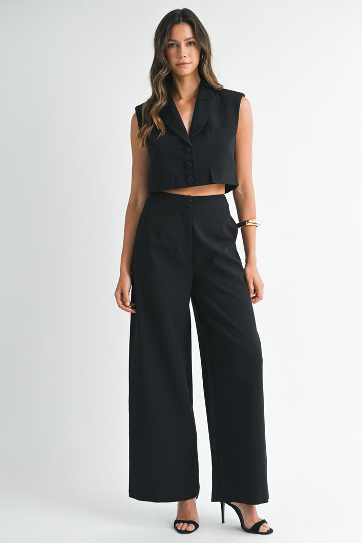 Model wearing a sleeveless black cropped vest and matching wide-leg pants with heels against a white background