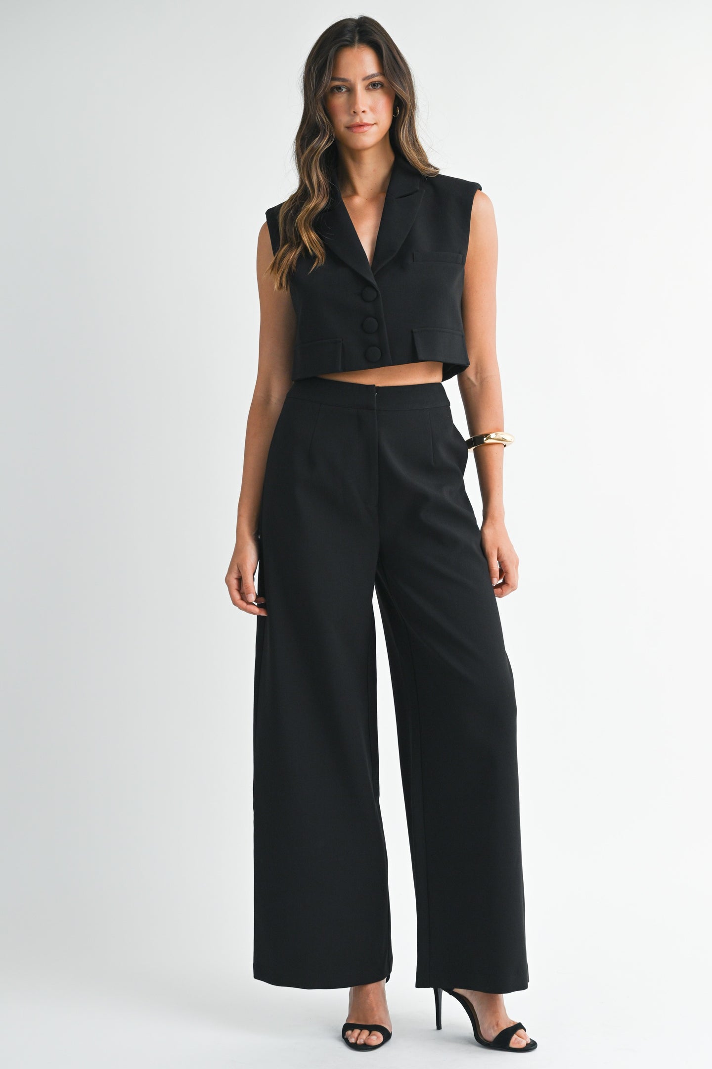 Model wearing a sleeveless black cropped vest and matching wide-leg pants with heels against a white background