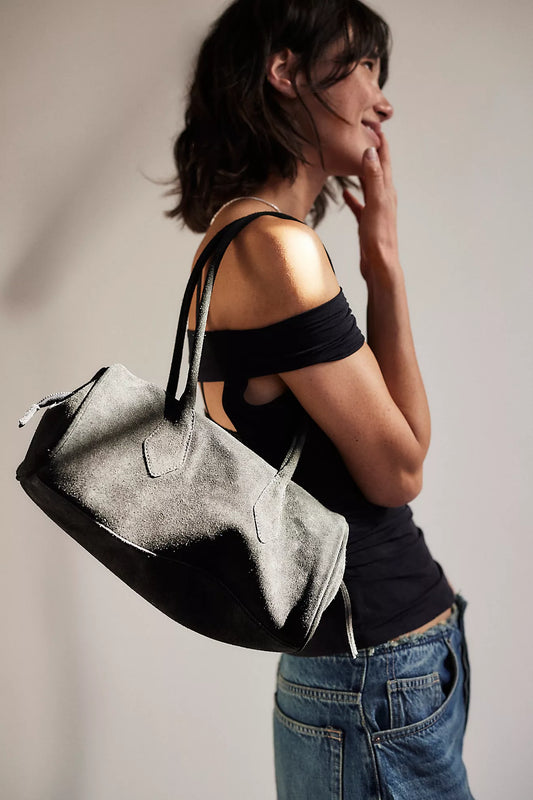 Model holding black suede tubular shoulder bag with dual straps and zip closure.