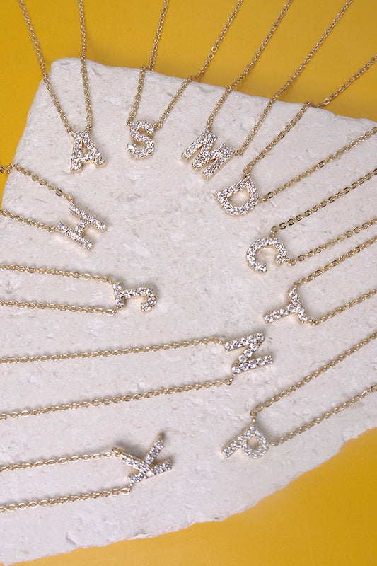 Gold necklaces with letter pendants on a white stand against a yellow background